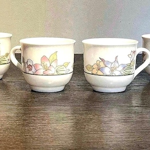 May include: Four white ceramic teacups with handles, each adorned with a floral design featuring pink, purple, and yellow flowers. A green band encircles the base of the floral pattern. The cups are arranged on a dark wooden surface.