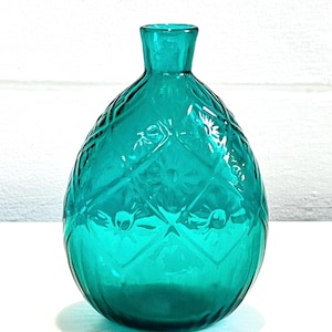 May include: A translucent teal glass vase with a rounded, bulbous body and a narrow neck. The exterior surface features a raised diamond pattern with embossed floral motifs centered within each diamond shape, creating a textured appearance.