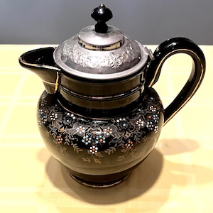 May include: Black ceramic teapot with a silver lid and handle. The teapot is decorated with a floral pattern in gold, white, and blue. The lid has an ornate design and a black knob. The spout is on the left side.