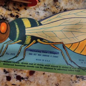 May include: A vintage cardboard toy bee with yellow and black stripes, blue wings, and a red head. The toy is attached to a string and hums when whirled around. The text on the toy reads "No. 205", "HUMS BEST WHEN CARDBOARD IS STRAIGHT", "MADE IN U.S.A", "WHIRL AROUND IN CIRCLES", and "DIRECTIONS: Grasp end of string, whirl around in circles or over the head. Different humming sounds can be obtained by adjusting rubber band".