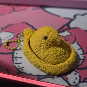 May include: A yellow Peep-shaped keychain with a textured surface and a small black eye. The keychain has a gold-colored chain attached. The background is a pink and white patterned surface.