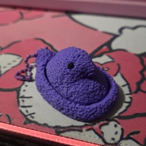 May include: A purple, textured keychain shaped like a Peep marshmallow chick. The chick has a small black eye and is attached to a chain with a small ring. The background is a pink surface with a cartoon design.