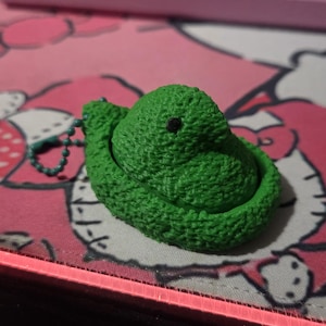 May include: A bright green, textured snake-shaped keychain charm with a small black eye. The charm is on a pink surface with a cartoon character design and a pink border.