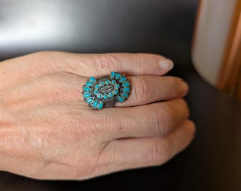 May include: A silver-toned ring with a central oval design and a cluster of small, bright turquoise stones. The ring has an ornate, detailed setting and is worn on a hand.