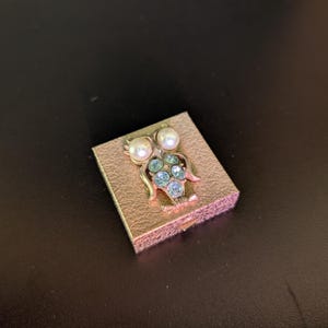 Op de afbeelding: Goudkleurige, rechthoekige broche met een uilontwerp. De uil heeft parelmoer ogen en een lichaam versierd met blauwe en heldere strass-steentjes. De broche heeft een getextureerd oppervlak en een vintage uitstraling.