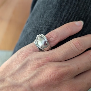 Puede incluir: Un anillo de plata hecho con una cuchara, con detalles florales ornamentados. El anillo se lleva en un dedo, mostrando el diseño de cubertería reciclada. La superficie pulida del anillo refleja la luz, resaltando su encanto vintage.