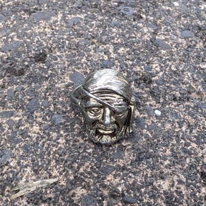 May include: A silver ring with a detailed pirate head design. The pirate is wearing a bandana and an eye patch.