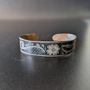 May include: A copper cuff bracelet with a black enamel band. The band features a floral and leaf design in a lighter colour. The bracelet has an open design and is likely adjustable. The cuff is a vintage or antique style.