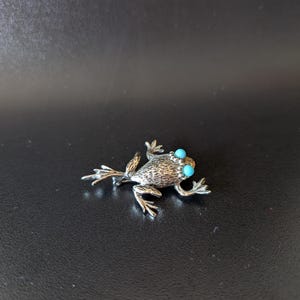 May include: A silver-toned frog brooch with turquoise blue eyes. The brooch has detailed texture on its body and legs, and is set against a dark background. The frog is a small, decorative accessory.