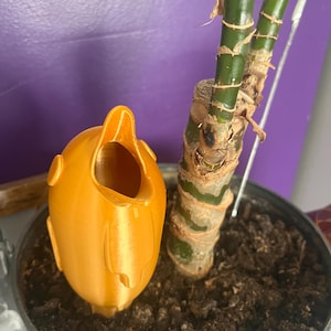 May include: A 3D printed orange plastic watering device shaped like a bird with its beak open. The device is placed in a pot with a green plant and brown soil.