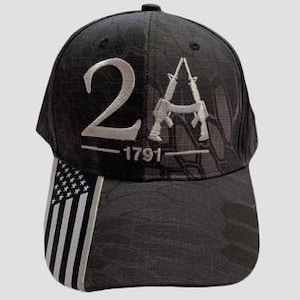 May include: Black baseball cap with the text "2A" and "1791" embroidered in white. The cap features a camouflage pattern and a graphic of two rifles. The left side of the cap displays a black and white American flag.