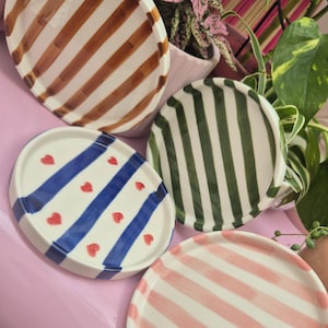 May include: A selection of decorative ceramic plates with striped and heart patterns. The plates display brown, green, pink, and blue stripes, and a white plate with blue stripes and red hearts. The plates are arranged on a pink surface.