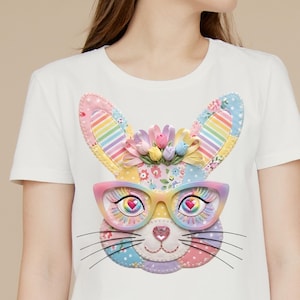 May include: White t-shirt featuring a colorful bunny face design. The bunny has rainbow-striped ears, a floral crown, and wears pink and yellow glasses with heart-shaped eyes. The design incorporates pastel colors and a patchwork style.