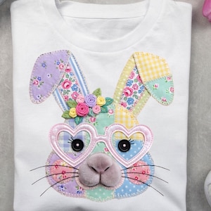 Pode incluir: Camiseta branca com um design de rosto de coelho em patchwork. O coelho tem óculos em forma de coração, detalhes florais, um nariz de pelúcia e olhos de botão. O design utiliza cores pastel e padrões florais.