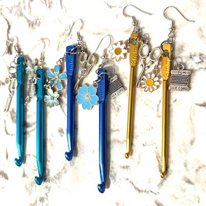 May include: Earrings featuring crochet hooks in teal, blue, and gold. Each earring has a hook charm with additional charms like flowers, scissors, and spools of thread. The hooks are metallic and have a shiny finish.