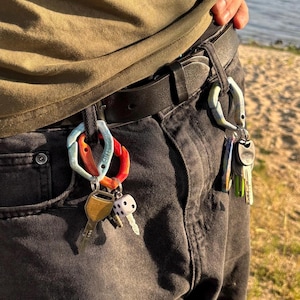 Könnte beinhalten: Nahaufnahme der Taille einer Person mit schwarzem Gürtel und dunkler Jeans. An den Gürtelschlaufen sind farbige Karabinerhaken befestigt, die Schlüssel und einen kleinen Würfel halten. Die Karabiner sind in Rot-, Orange-, Blau- und Weißtönen gehalten.