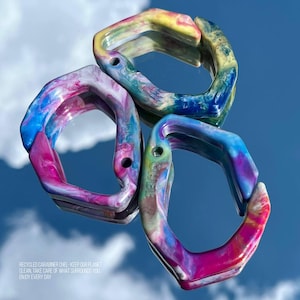 Puede incluir: Tres mosquetones reciclados multicolores con efecto marmolado. Los clips muestran tonos de rosa, azul, amarillo y verde. La palabra "CHEL" es visible en cada clip. El texto "RECYCLED CARABINER CHEL - KEEP OUR PLANET CLEAN, TAKE CARE OF WHAT SURROUNDS YOU, ENJOY EVERY DAY" está en la parte inferior.