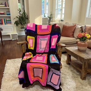 Puede incluir: Una colorida manta de ganchillo hecha a mano, sobre una silla de madera. La manta presenta un diseño de patchwork con cuadrados rosas, naranjas, amarillos y azules sobre un fondo oscuro. La silla está sobre una alfombra mullida de color blanco roto.