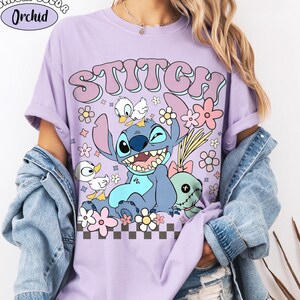 Könnte beinhalten: Ein orchideenfarbenes T-Shirt mit einer Grafik der Figur Stitch, zwei kleinen weißen Enten und einer Puppe. Der Text STITCH ist in großen violetten Buchstaben über der Figur gedruckt. Ein kleines Etikett zeigt COMFORT COLOR Orchid.