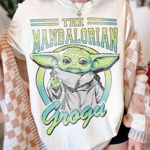 May include: A cream-colored t-shirt featuring a graphic print of the character Grogu from The Mandalorian. The design includes the text THE MANDALORIAN in green and blue gradient lettering above the character and the name Grogu in blue below.