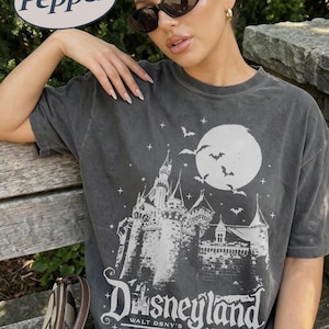 K&ouml;nnte beinhalten: Dunkelgraues T-Shirt mit einem Disneyland-Motiv. Das Design zeigt ein Schloss, Flederm&auml;use und einen Vollmond. Der Text lautet "Disneyland Walt DSNY's Anaheim" und "I survived the happiest place on earth."