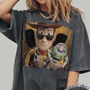 K&ouml;nnte beinhalten: Ein anthrazitfarbenes Kurzarm-T-Shirt mit einem Grafikdruck von Woody mit Sonnenbrille, der Buzz Lightyear h&auml;lt. Das T-Shirt hat eine verwaschene Vintage-Optik. Der Text Comfort Colors Pepper ist unten rechts auf das T-Shirt aufgedruckt.