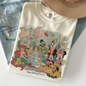 May include: Cream-colored t-shirt featuring a colorful graphic of Disney characters, including Dumbo, Ariel, Mickey Mouse, and Minnie Mouse. The text "Magic Kingdom Walt Disney World" is printed below the characters. The shirt is folded on top of blue jeans.
