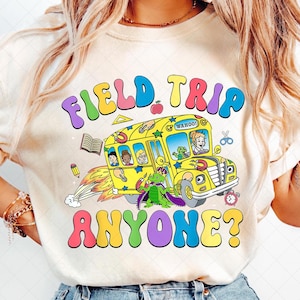 Può includere: Una maglietta bianca con una grafica colorata di un autobus scolastico con il testo "FIELD TRIP ANYONE?" in lettere arcobaleno.