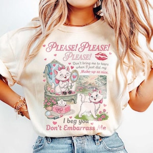 Könnte beinhalten: Ein cremefarbenes T-Shirt mit einer Grafik von zwei weißen Katzen mit rosa Schleifen. Der Text lautet: Please! Please! Please! Don't bring me to tears when I just did my Make-up so nice. I beg you Don't Embarrass Me, mit rosa Herzen und Lippen.