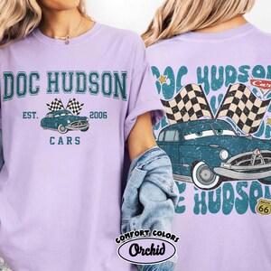 May include: A light purple t-shirt featuring a graphic of a vintage blue car with checkered flags and the text "DOC HUDSON CARS EST. 2006". The back of the shirt also has the same car graphic and text.