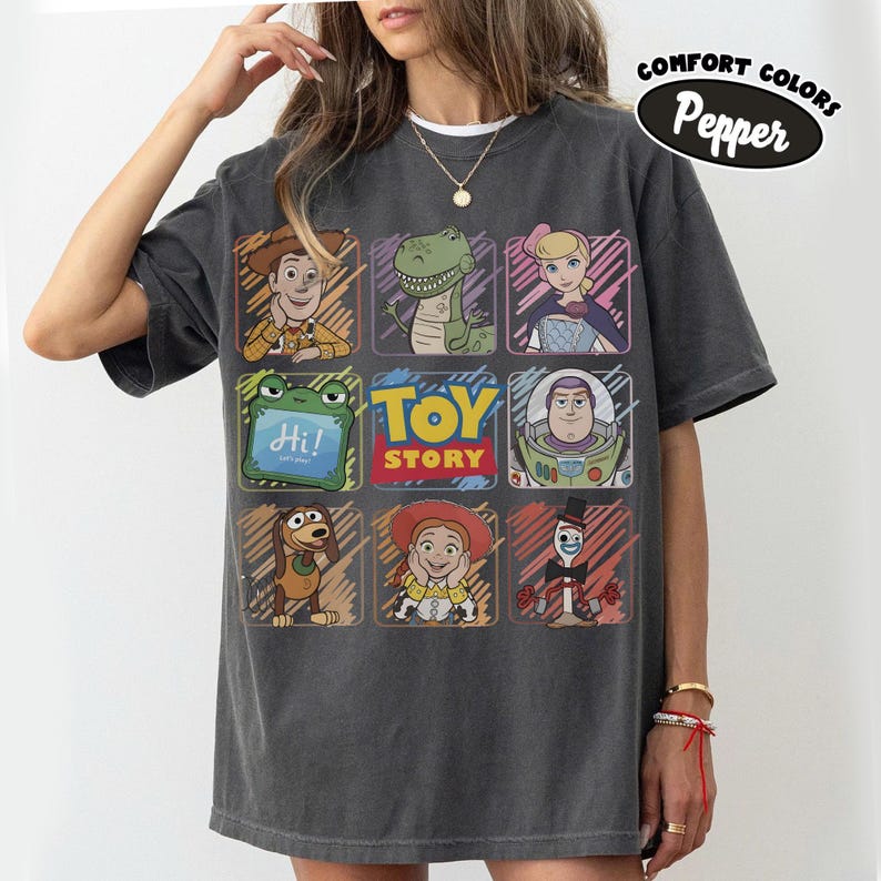 May include: Dark grey comfort colours t-shirt featuring a grid of Toy Story characters. The shirt includes Woody, Rex, Bo Peep, Buzz Lightyear, Slinky Dog, Jessie, and Mr. Pricklepants. The text "TOY STORY" is also displayed. The shirt has the text "Comfort Colors Pepper".