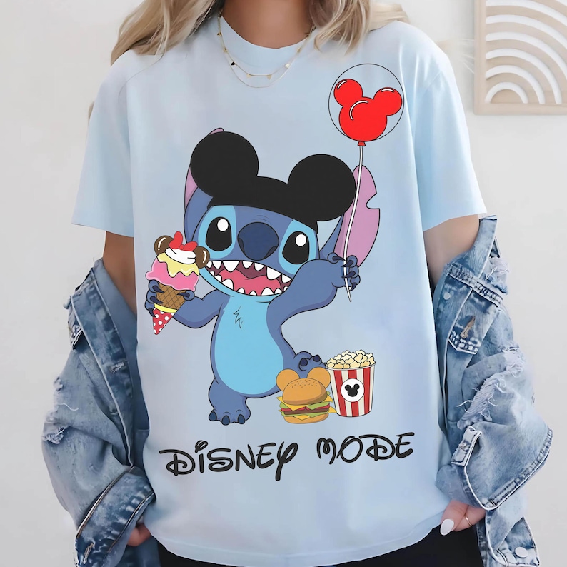 Puede incluir: Camiseta azul claro con un personaje de dibujos animados con un sombrero negro de Mickey Mouse, sosteniendo un helado y un globo rojo. La camiseta tambi&eacute;n tiene un gr&aacute;fico de hamburguesa y palomitas de ma&iacute;z, con las palabras "DISNEY MODE" en negro.