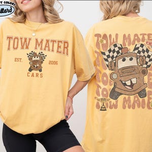 May include: Mustard-colored t-shirt featuring the text "TOW MATER" above a cartoon image of Tow Mater holding checkered flags. The back of the shirt repeats the design. The shirt also has the text "Cars" and "Est. 2006".