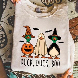 Könnte beinhalten: Beiges Sweatshirt mit Halloween-Motiv, das drei Comic-Enten in Kostümen zeigt. Eine ist ein Kürbis, eine andere ein Geist und die dritte eine Hexe. Der Schriftzug "DUCK, DUCK, BOO" ist darunter gedruckt, mit Fledermaus-Silhouetten.