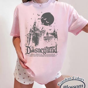K&ouml;nnte beinhalten: Hellrosa T-Shirt mit einem grauen Motiv eines Schlosses, Mondes und Flederm&auml;usen, mit dem Wort "Disneyland" und "Anaheim" darunter. Der Text "I survived the happiest place on Earth" ist ebenfalls aufgedruckt. Das T-Shirt ist ein Comfort Colors Blossom.