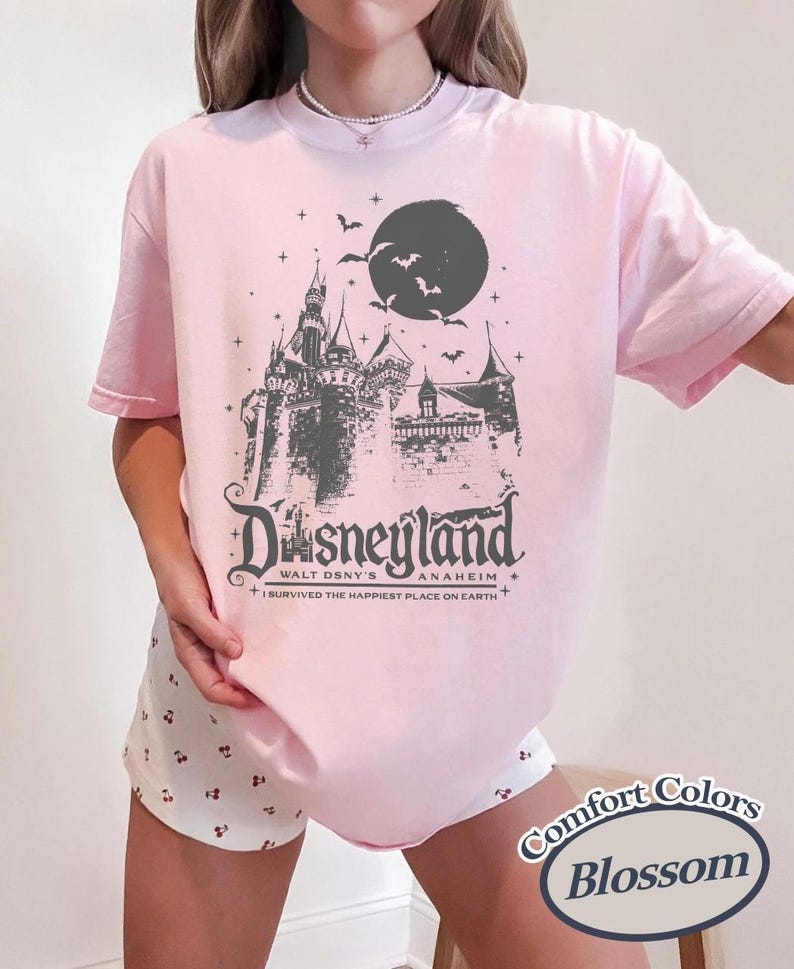 K&ouml;nnte beinhalten: Hellrosa T-Shirt mit einem grauen Motiv eines Schlosses, Mondes und Flederm&auml;usen, mit dem Wort "Disneyland" und "Anaheim" darunter. Der Text "I survived the happiest place on Earth" ist ebenfalls aufgedruckt. Das T-Shirt ist ein Comfort Colors Blossom.