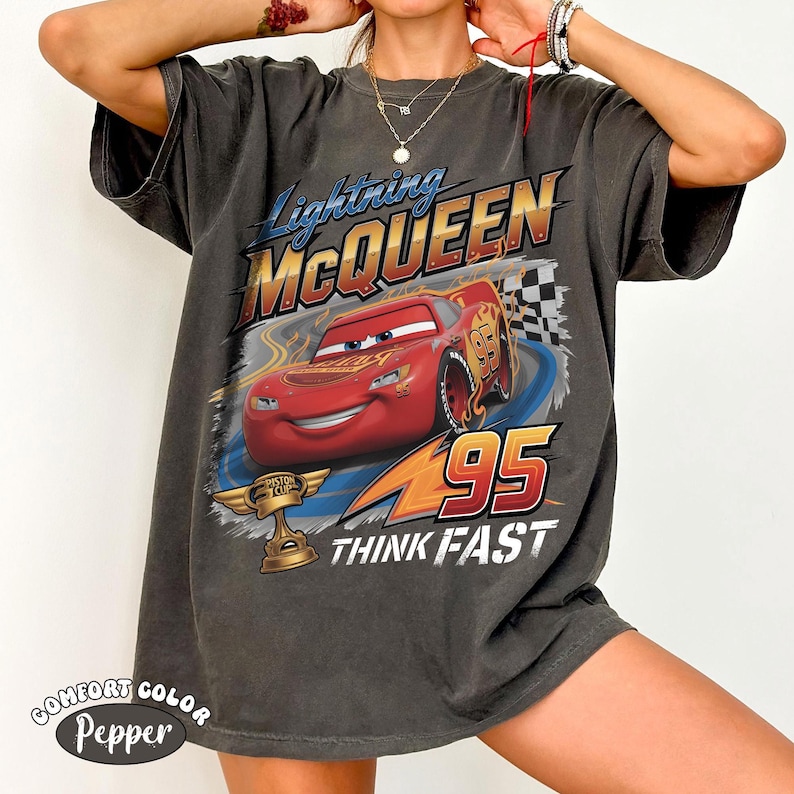 May include: A charcoal gray short-sleeve t-shirt featuring a graphic of the red race car Lightning McQueen. The design includes the text Lightning McQUEEN, the number 95, a Piston Cup trophy, and the words THINK FAST in white lettering.