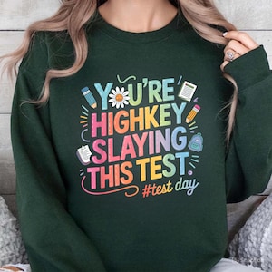 Könnte beinhalten: Grüner Pullover mit einer Regenbogengrafik, auf der "You're Highkey Slaying This Test" mit Illustrationen von Blumen, Bleistiften, Notizbüchern und Rucksäcken steht. Der Text "#test day" befindet sich unter der Grafik.