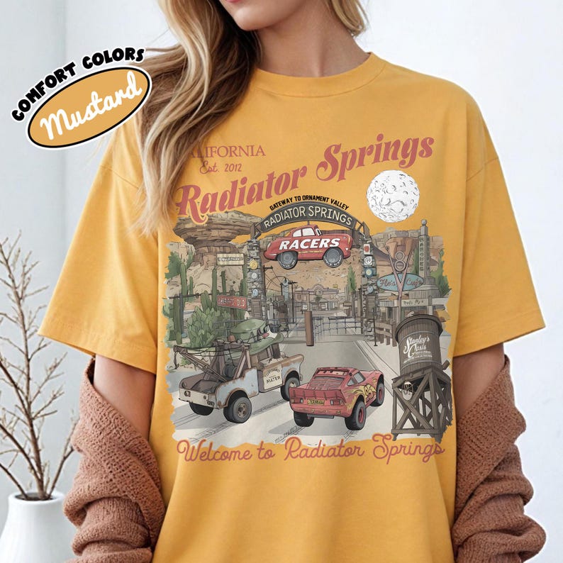 May include: Mustard-colored t-shirt featuring a graphic of Radiator Springs, with text that reads "Radiator Springs" and "Welcome to Radiator Springs." The design includes images of cars and a water tower. The shirt has short sleeves.