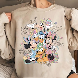 Könnte beinhalten: Beiges Sweatshirt mit einem farbenfrohen Aufdruck von Disney-Figuren, darunter Micky Maus, Minnie Maus, Donald Duck und Goofy, mit Ostermotiven. Das Design beinhaltet Unterschriften der Charaktere und florale Akzente.