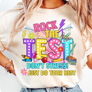 Könnte beinhalten: Weißes T-Shirt mit einer bunten Grafik, auf der "Rock the Test! Don't Stress! Just Do Your Best" steht. Die Grafik enthält eine Vielzahl von Schulutensilien wie Bleistifte, Büroklammern und einen roten Apfel.