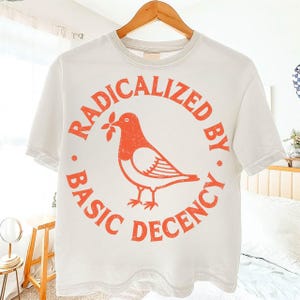 Könnte beinhalten: Weißes T-Shirt mit einem orangefarbenen, verblassten Aufdruck eines Vogels, der einen Zweig im Schnabel hält, umgeben von den Worten "RADICALIZED BY . BASIC DECENCY." Das Shirt hängt an einem Holzkleiderbügel.