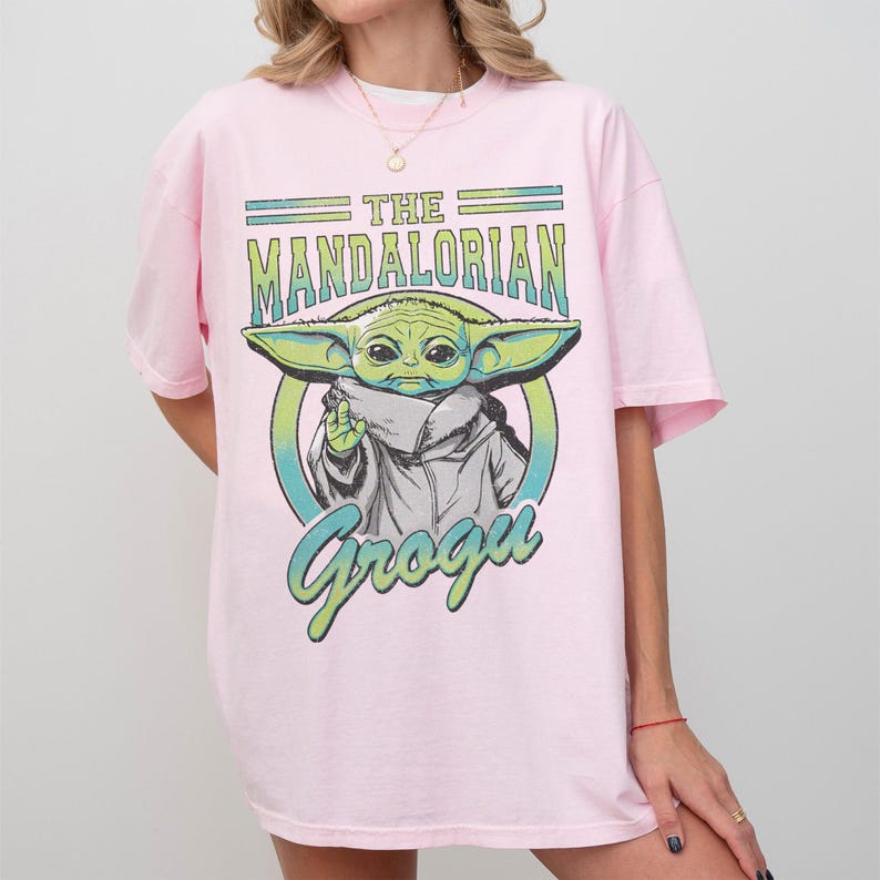 May include: A light pink short-sleeve t-shirt featuring a graphic print of Grogu from The Mandalorian. The design includes the text THE MANDALORIAN and Grogu in a distressed green and blue font, centered over a circular background element.