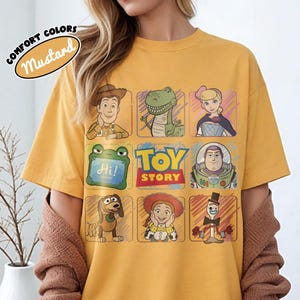 May include: Mustard-coloured t-shirt with a grid of Toy Story character illustrations. The shirt features Woody, Rex, Bo Peep, Buzz Lightyear, Slinky Dog, Jessie, and Mr. Pricklepants. The words "TOY STORY" are in the centre.