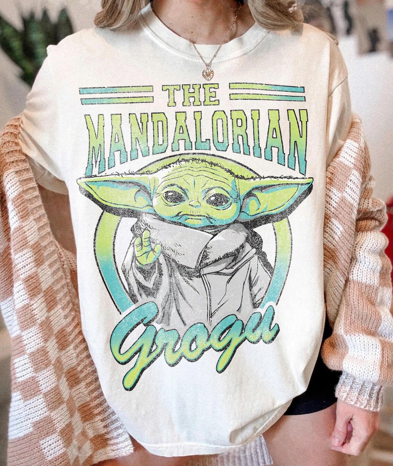 May include: A cream-colored t-shirt featuring a graphic print of the character Grogu from The Mandalorian. The design includes the text THE MANDALORIAN in green and blue gradient lettering above the character and the name Grogu in blue below.