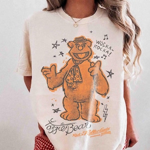 Könnte beinhalten: Ein cremefarbenes T-Shirt mit einer Grafik eines braunen Bären, der einen Hut und ein gepunktetes Halstuch trägt. Der Aufdruck enthält den Text WOCKA-ROCKA!, Fozzie Bear und Rock'n Roller Coaster, umgeben von Sternen, Noten und Blitzen.
