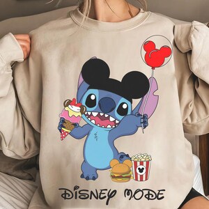Puede incluir: Sudadera beige con un personaje de dibujos animados azul con orejas negras, sosteniendo un helado y un globo rojo de Mickey Mouse. El personaje est&aacute; rodeado de alimentos y las palabras "Disney Mode".