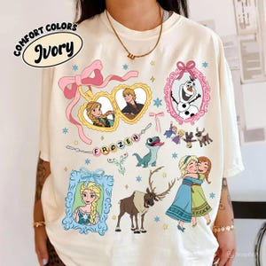 Könnte beinhalten: Elfenbeinfarbenes Comfort Colors T-Shirt mit farbenfrohen Grafiken aus dem Film Frozen. Das Design umfasst Bilder von Anna, Elsa, Olaf, Kristoff, Sven und anderen Charakteren. Das Wort "FROZEN" ist ebenfalls abgebildet.