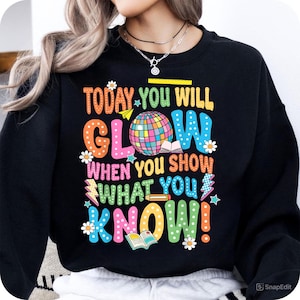 May include: Black sweatshirt with a colorful graphic that says "Today you will glow when you show what you know!" The graphic includes a globe, a book, a pencil, and stars.