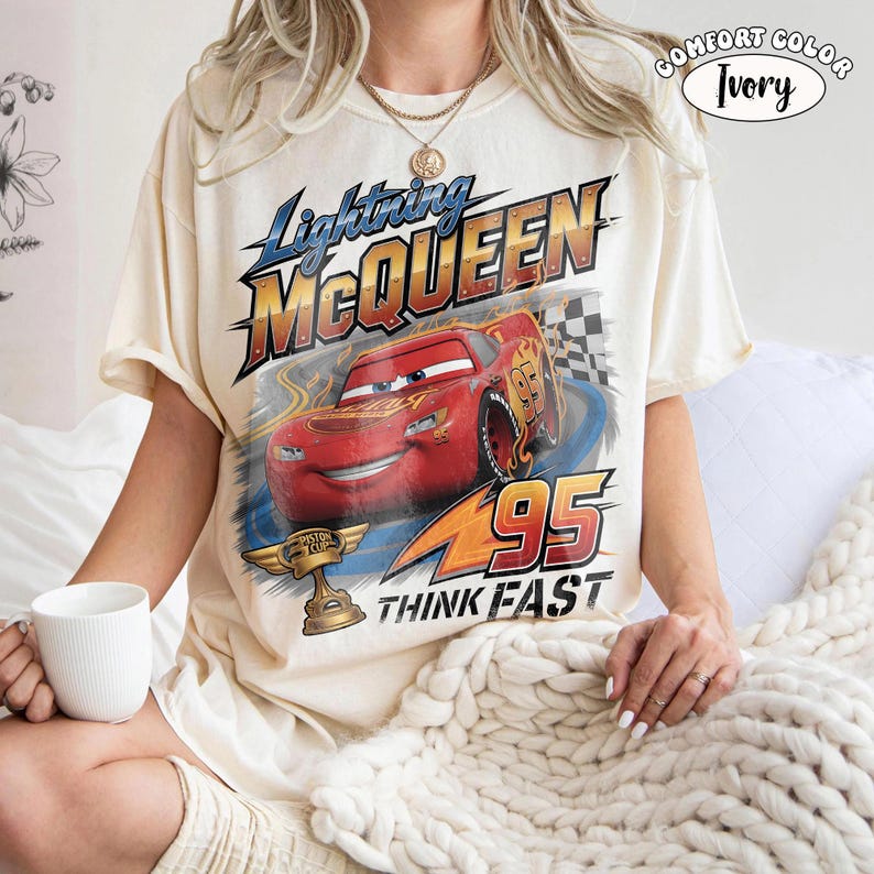 May include: An ivory short-sleeve t-shirt featuring a graphic of the red race car Lightning McQueen. The design includes the text Lightning McQueen, the number 95, a gold Piston Cup trophy, and the words Think Fast in a bold font.